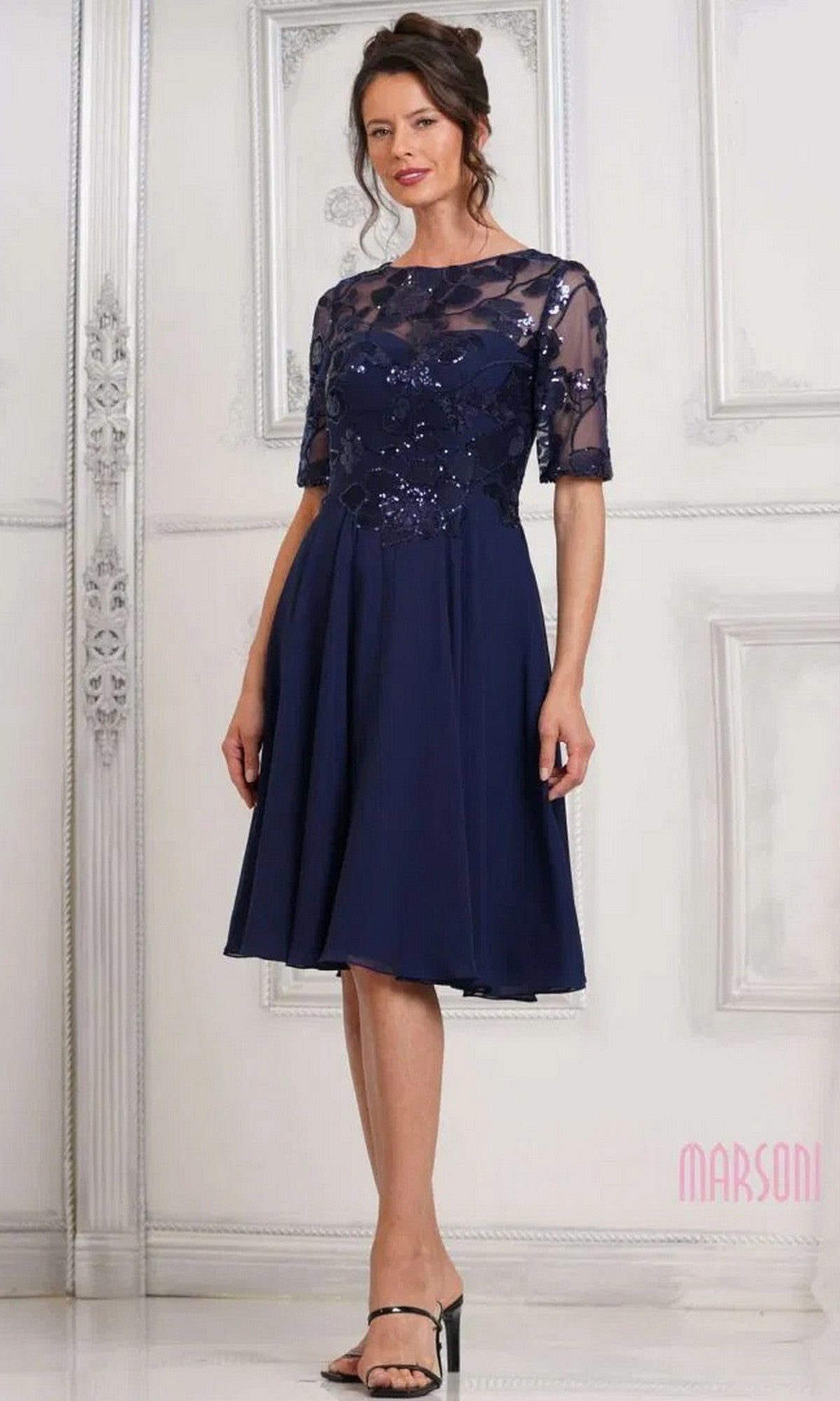 Woman in the Marsoni M286S semi-formal knee-length navy dress, featuring floral sequin lace details and sheer sleeves, standing indoors against an ornate light-colored wall.